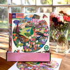 Jigsaw puzzle box with a colorful design of birds and flowers on a wooden table with a vase of flowers in the background.