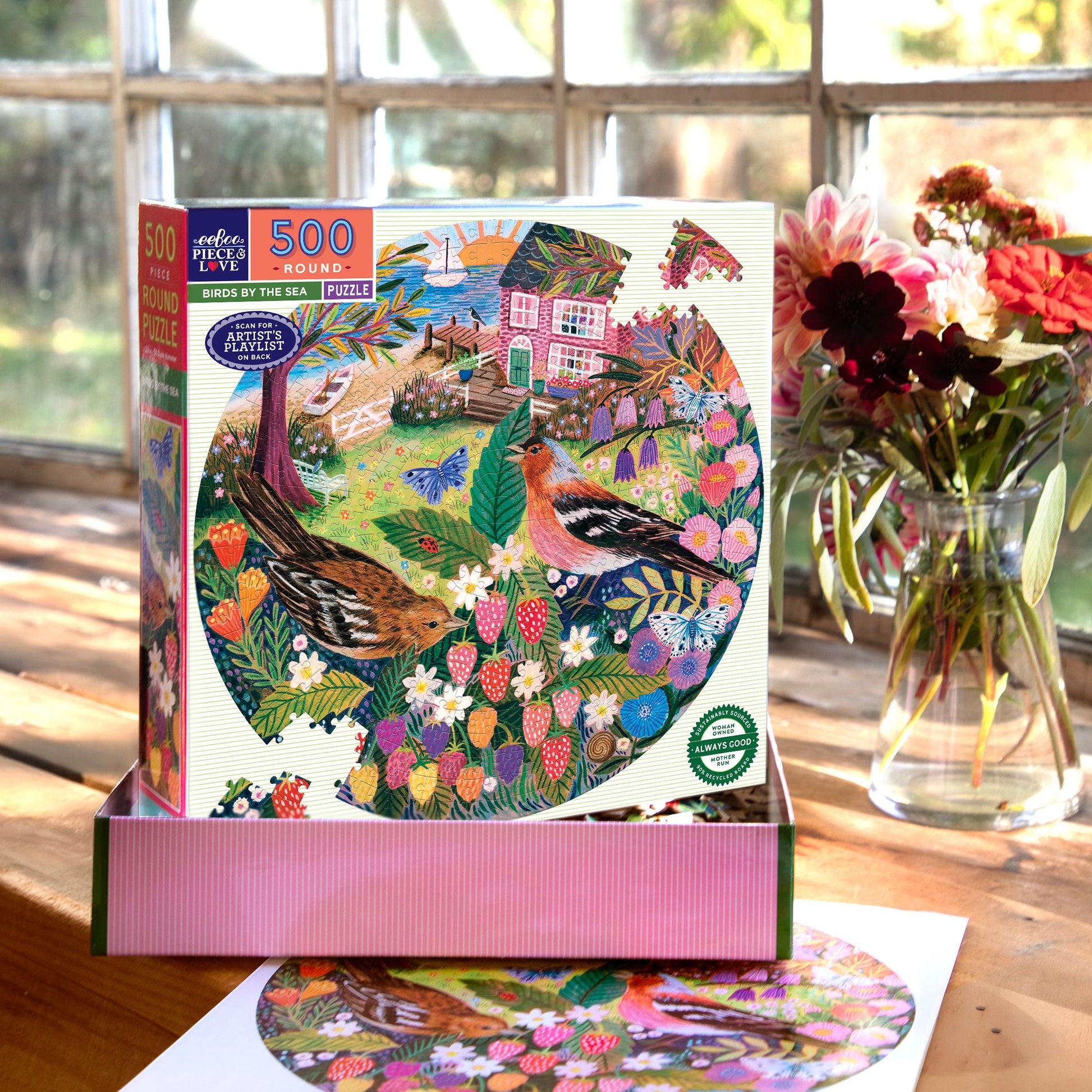 Jigsaw puzzle box with a colorful design of birds and flowers on a wooden table with a vase of flowers in the background.