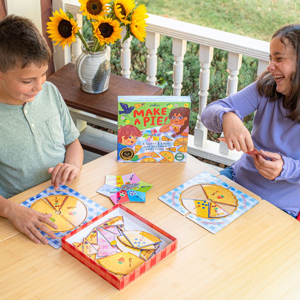 Two children playing with a 'Make a Pie!' educational game on a wooden table.