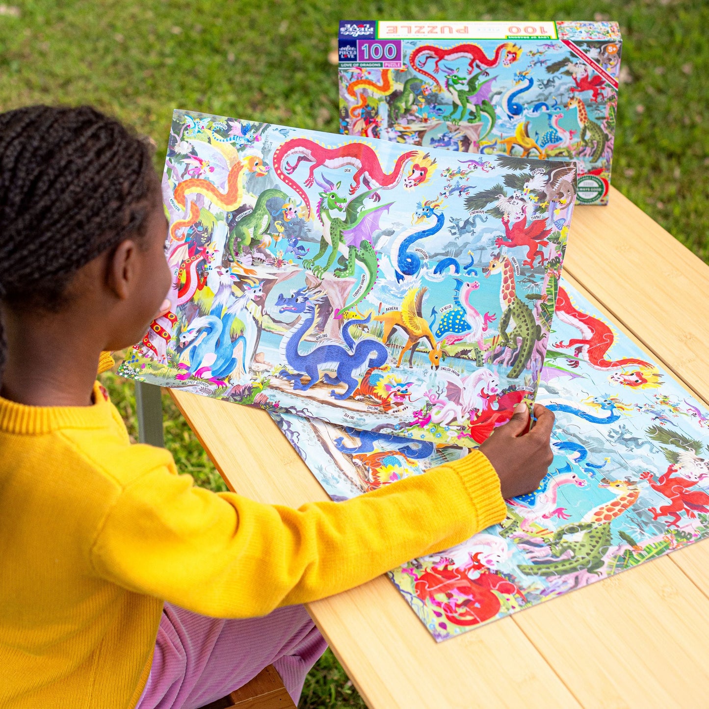 Child playing with a colorful dragon puzzle on a wooden table outdoors. Love of Dragons 100 Piece Puzzle