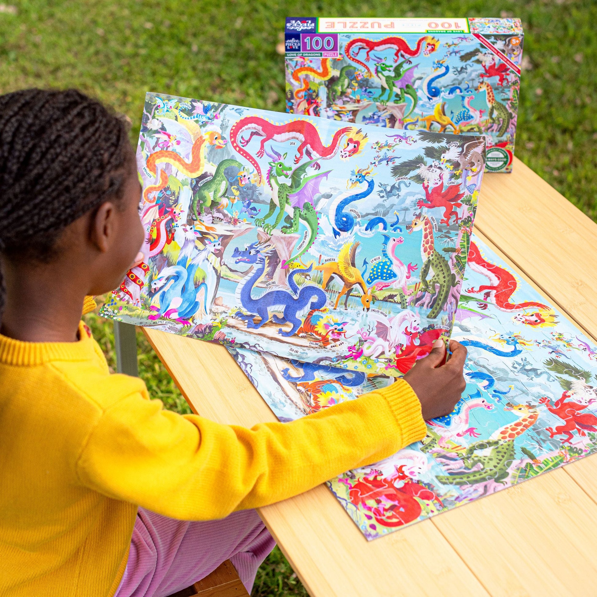 Child playing with a colorful dragon puzzle on a wooden table outdoors. Love of Dragons 100 Piece Puzzle