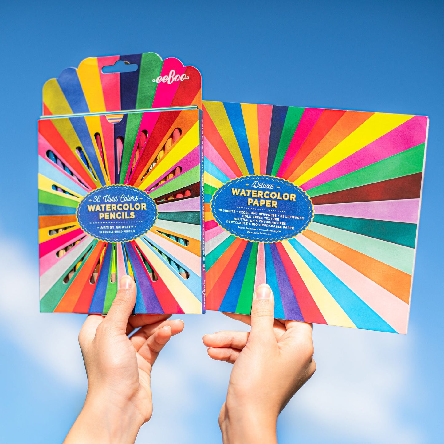 Two packages of watercolor pencils and paper held against a blue sky. Rainbow Watercolor Pad
