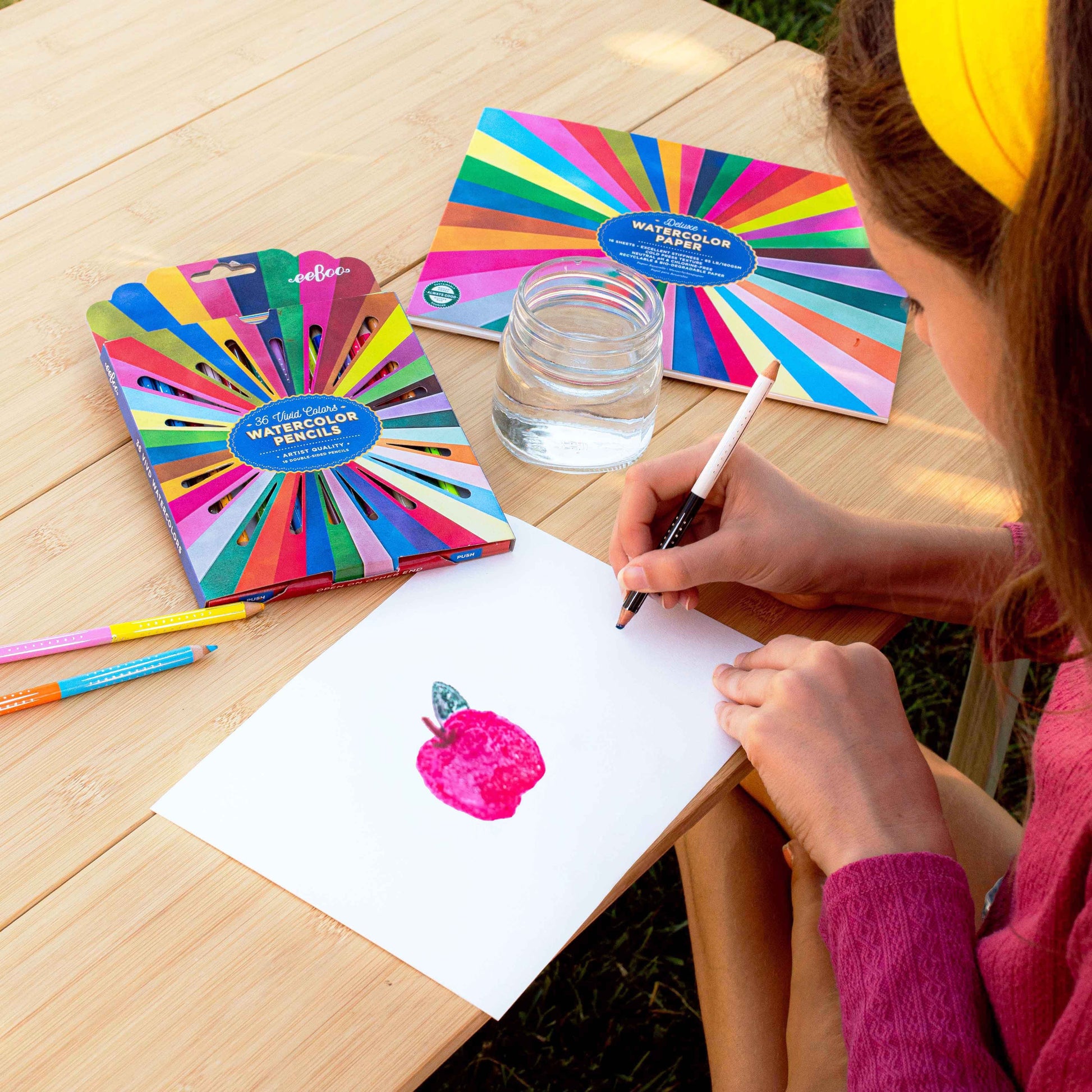 Person drawing with watercolor pencils on a wooden table with colorful pencil sets. Rainbow Watercolor Pad