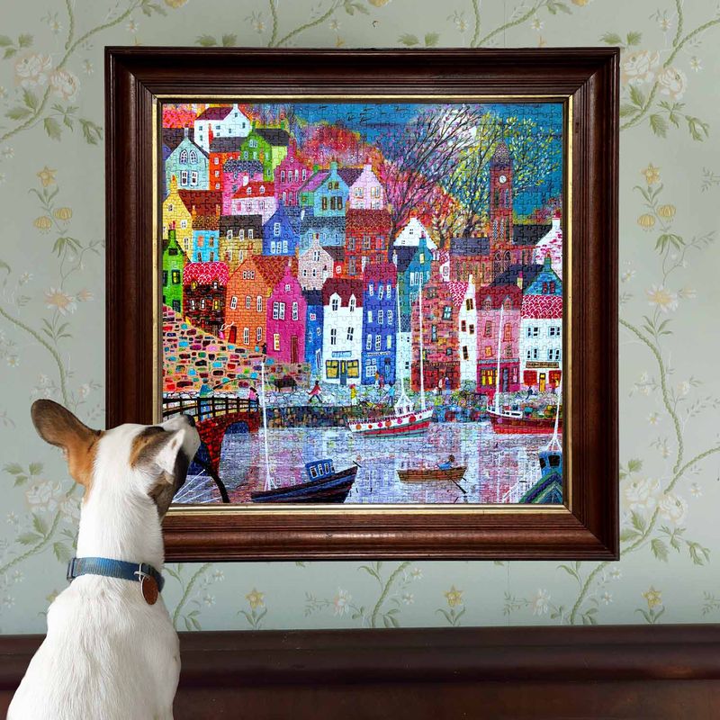 Dog looking at a colorful painting of a town by a river on a floral wallpapered wall.