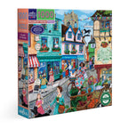Jigsaw puzzle box featuring a colorful village scene with shops and people. Village by the Sea 1000 Piece Puzzle